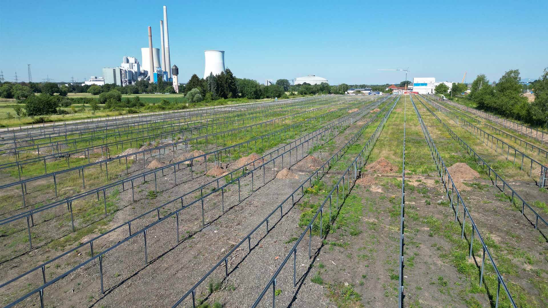 Baufeld mit montierten Metallgestellen für eine zukünftige Solaranlage, im Hintergrund ein Industriegebiet.