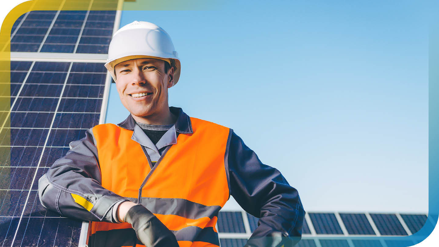 Techniker mit Schutzhelm und Warnweste steht vor Solarmodulen und lächelt in die Kamera.